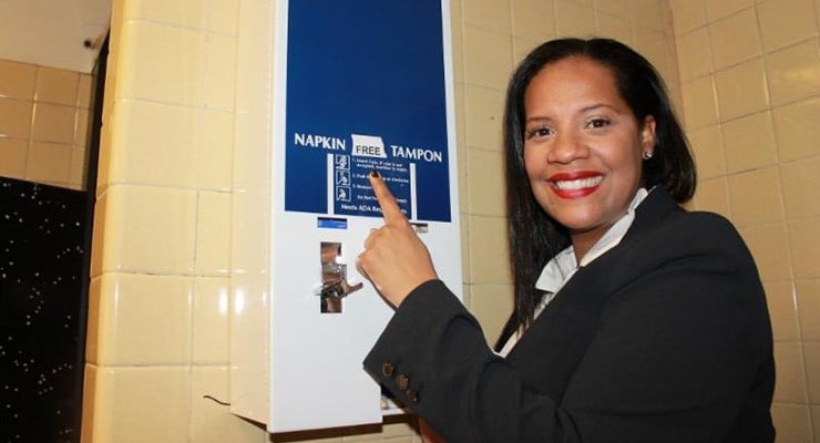 Woman pointing to the text 'free' on a sanitary napkin dispenser in a bathroom