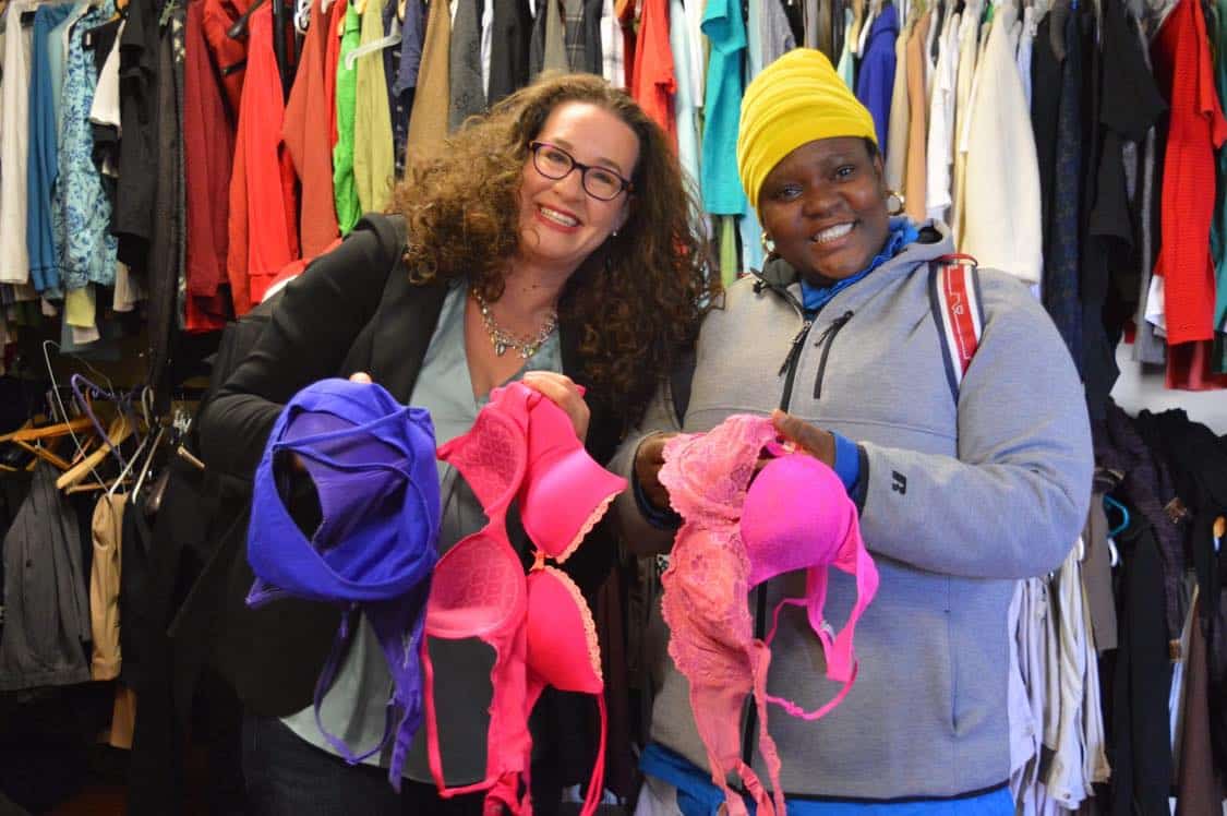 Two women standing side by side, holding bras. The woman on the right, Dana Marlowe, has brought bras to a homeless shelter so that women experiencing homelessness- the woman on the left- can receive them.